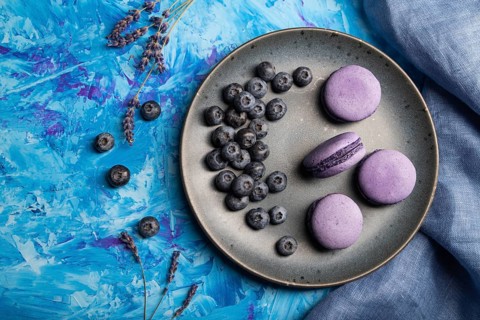 Purple macarons or macaroons cakes with blueberries on ceramic plate on a blue concrete background