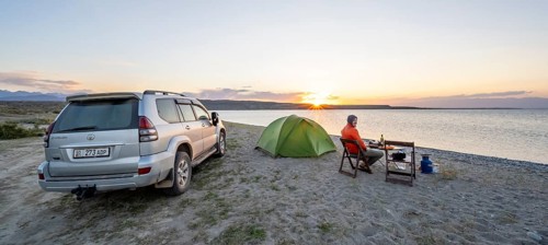 Camping at the lakeside, young man sitting next to tent on camping chair, having dinner, off-road