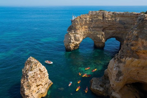 Stone arches and rocks jut out of the clear blue sea, kayaks and a small boat glide across the