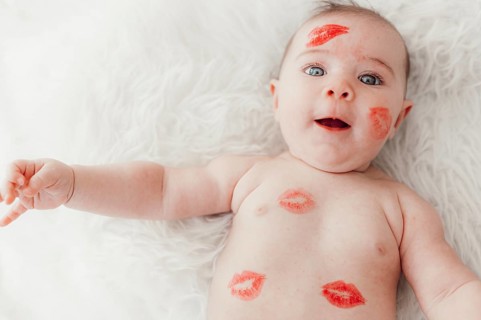 A smiling baby with red lipstick kisses on her face and body lies on a white blanket and looks into