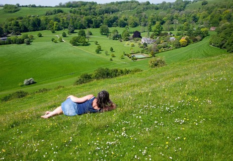 Woman lying on chalk grassland looking into deep green valley containing Rainscombe House, Oare