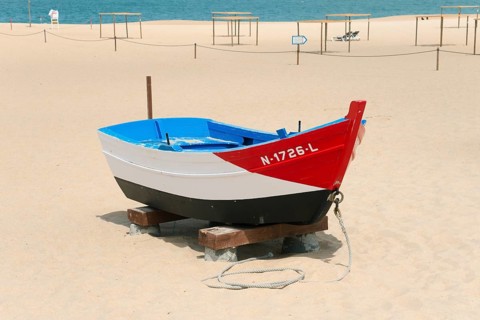 A small red, blue and white boat on a sandy beach, tied up with a rope, Praia da Nazare beach,