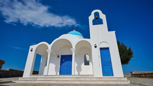 White church with blue doors and a bell in front of a blue sky and tree, Church of Profitis Ilias,