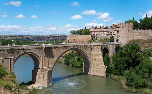 Historic stone bridge over river in rural setting with neighbouring old buildings, Puente de San