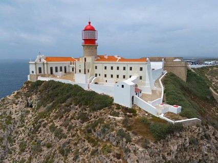 A red lighthouse and white buildings stand on a cliff, surrounded by vegetation and the ocean,