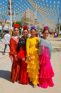 Four woman in bright flamenco dresses and flowers in their hair walk along a festival street under