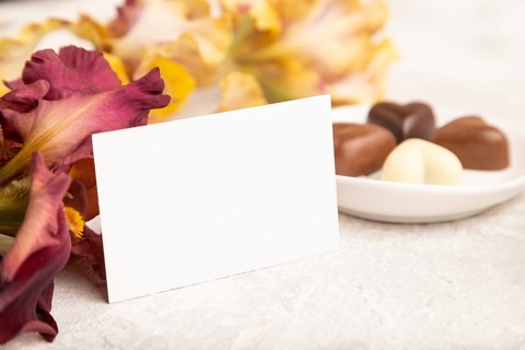 White business card with chocolate candies and iris flowers on gray concrete background. side view,