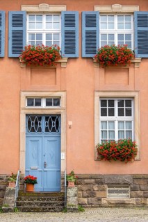Picture of a traditional house with blue front door and shutters, decorated with red geraniums in