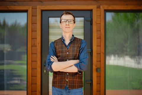 Middle-aged man in blue shirt and brown vest stands confidently in front of modern wooden house