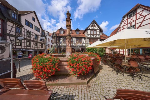 Market fountain with geraniums and outdoor area of a restaurant under a blue sky with cumulus