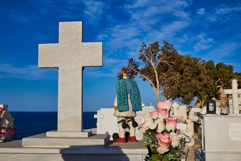 A cemetery by the sea with gravestones and crosses. In the foreground are flowers and a figure with