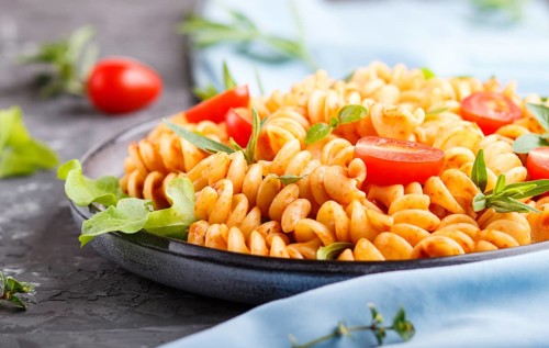 Fusilli pasta with tomato sauce, cherry tomatoes, lettuce and herbs on a black concrete background.