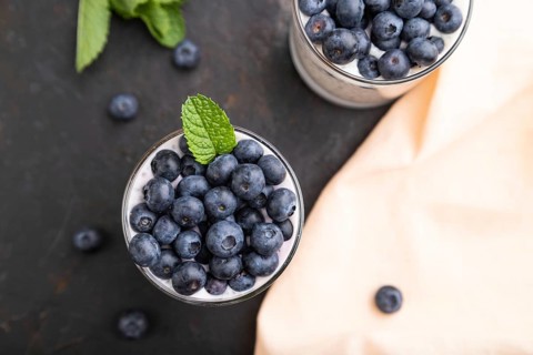 Yogurt with blueberry and chia in glass on black concrete background and orange linen textile. top