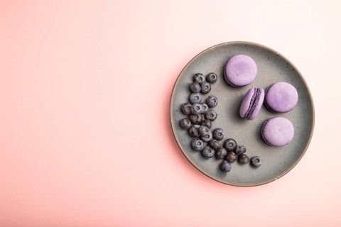 Purple macarons or macaroons cakes with blueberries on ceramic plate on a pastel pink background.
