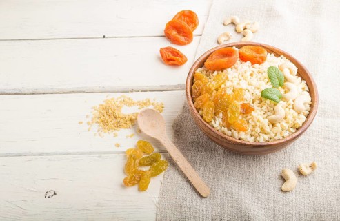 Bulgur porridge with dried apricots, raisins and cashew in wooden bowl on a white wooden background