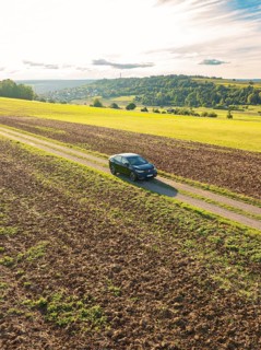 Black car driving on a gravelled country lane through a wide, rural landscape under a cloudy sky,