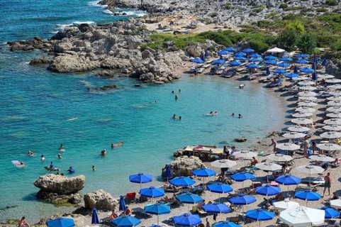 Well frequented beach with many blue parasols and swimmers in the emerald green sea under the