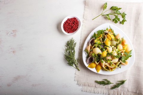 Conchiglie colored pasta with fresh greengrocery on a linen tablecloth on white wooden background.