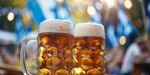 Banner with large beer mugs with blue and white Oktoberfest festival flags in background.