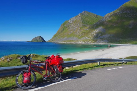 Bicycle with red bags on a road along a beach with turquoise water, in the middle of mountains and