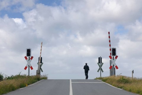 A man walks along an empty road approaching a railway crossing with traffic lights under an