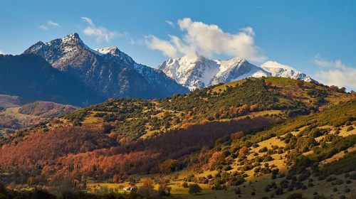 Autumn mountain landscape with snow-covered peaks under a cloudy blue sky, autumn foliage