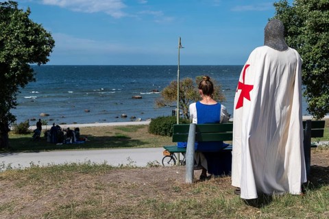 A knight with a red cross on a white coat and armour stands behind a woman sitting on a bench with