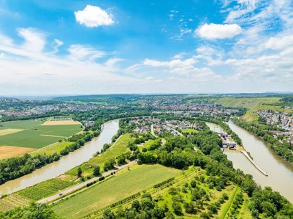 Hilly landscape with wide fields and a meandering river under a blue sky, with a small village on