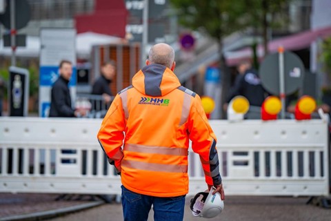 A worker in an orange safety waistcoat with a helmet stands in front of a barrier on the road,