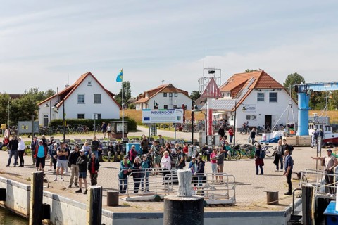 People gather at the harbour next to buildings and bicycles, a flag in the background, Rügen,