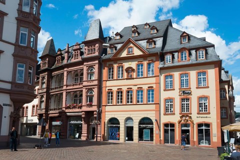 Multi-storey historic buildings with red facades and shutters surround a lively town square,