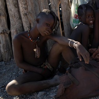 Himba girl with braided pigtails sitting in the shade near a hut, near Opuwo, Kaokoveld, Kunene,