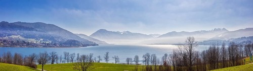A picturesque lake surrounded by mountains and trees under a blue, slightly misty sky, Tegernsee in