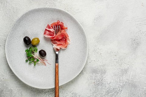 Prosciutto on a fork, top view, olives and micro-greens on a plate, light background, no people