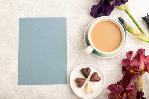 Blue paper sheet with cup of cioffee, chocolate candies, purple and burgundy iris flowers on gray