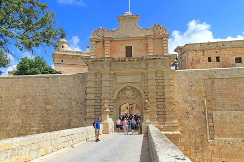 Tourists walking over moat bridge through entrance gateway of medieval city of Mdina, Malta, Europe