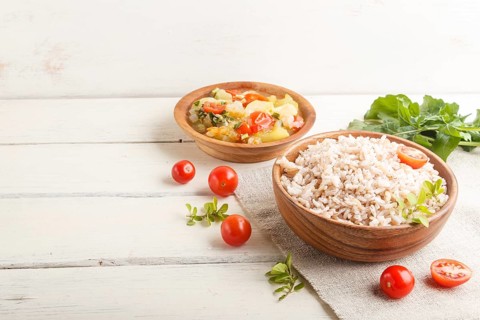 Unpolished rice porridge with stewed vegetables and oregano in wooden bowl on a white wooden
