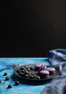 Purple macarons or macaroons cakes with blueberries on ceramic plate on a blue and black background