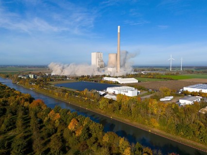 A coal-fired power station with smoking chimneys in a rural setting, surrounded by wind turbines