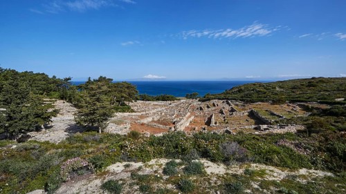 Expansive landscape of ruins overlooking the sea, surrounded by vegetation under a clear blue sky,