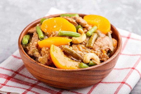 Fried pork with peaches, cashew and green beans in a wooden bowl on a gray concrete background.