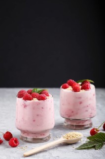 Yoghurt with raspberry and sesame in a glass and wooden spoon on gray and black background. side