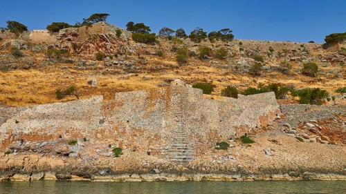 Old stone wall on rocky cliff with dry vegetation and calm sea water in the foreground, Venetian