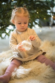 A toddler holds a glittering Christmas bauble in front of a Christmas tree in a cosy, wintry