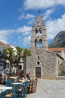 Traditional stone church with bell tower next to a café with terrace under a clear blue sky