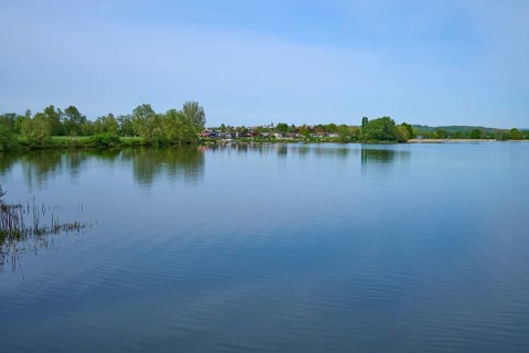 Quiet lake with trees on the shore, in the background a campsite, leisure area, under blue sky,