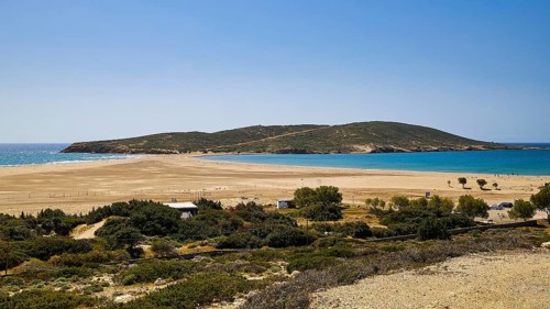 A quiet beach with sand dunes and a lush green island in the background under a bright blue sky,