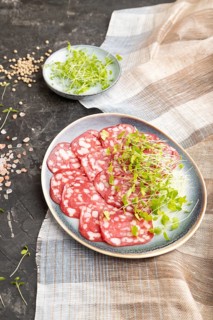 Slices of smoked cervelat salami sausage with spinach microgreen, salt and pepper on black concrete
