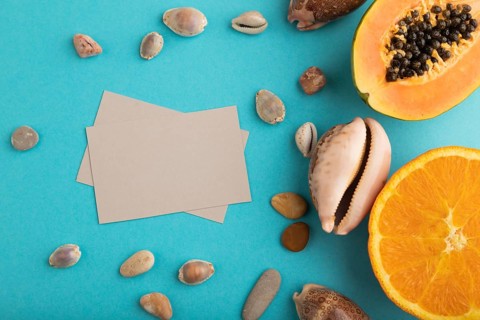 Gray paper business card with ripe cut papaya, orange, seashells on blue pastel pastel background.