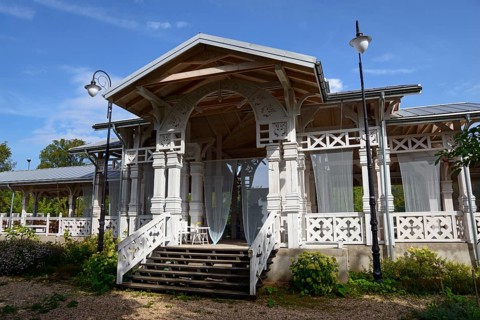An elegant wooden veranda with white columns and stairs, surrounded by plants under a blue sky, Old